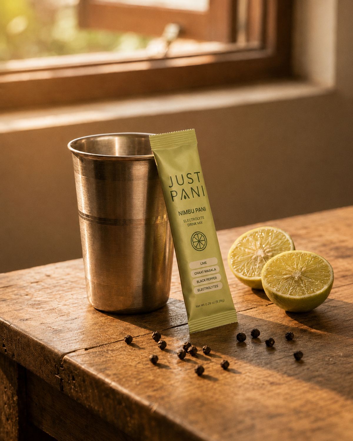 A steel tumbler and a JUSTPANI packet on a wooden table beside cut limes and black peppercorns, lit by a window