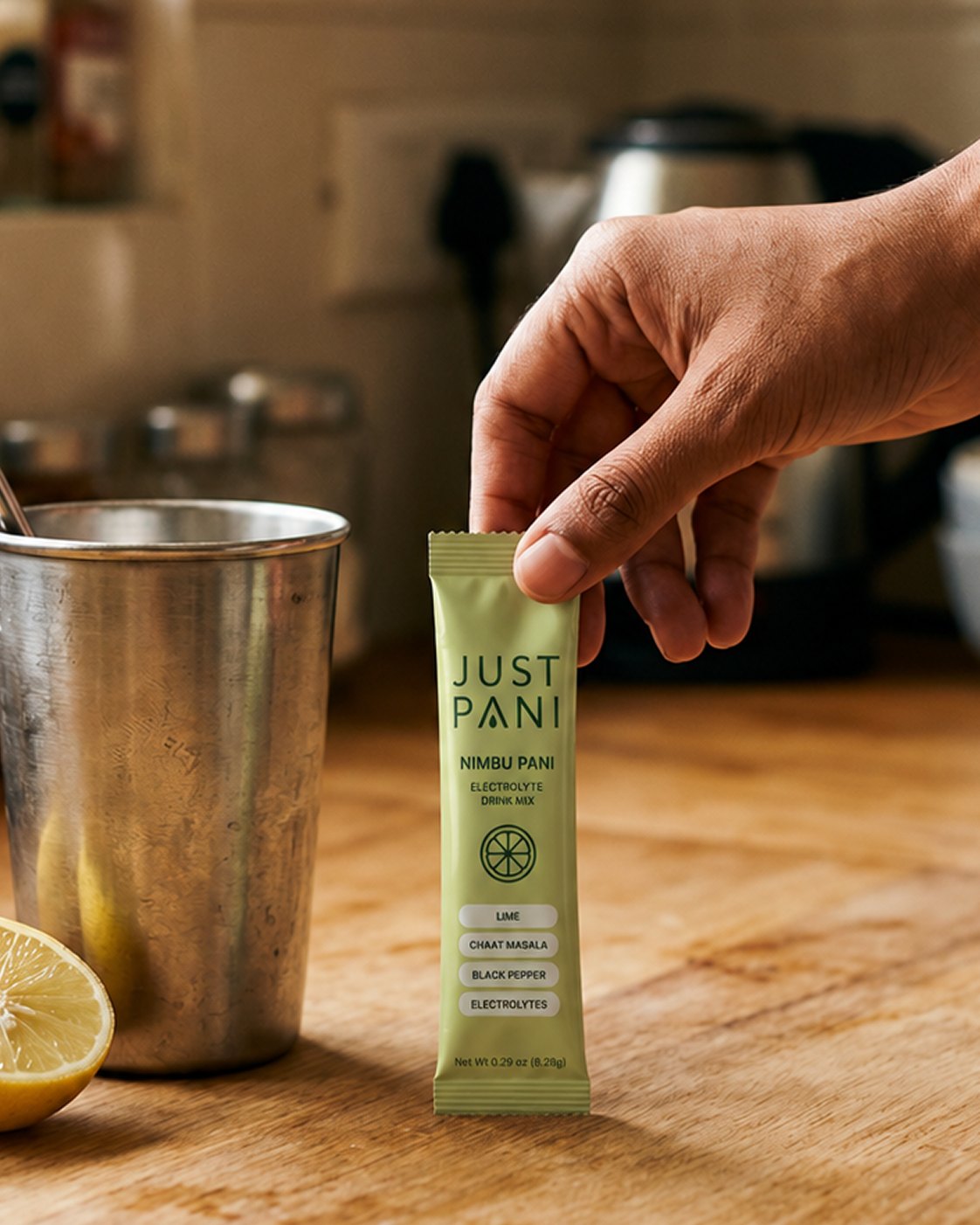 A hand holding a JUSTPANI Nimbu Pani packet over a wooden kitchen counter, beside a steel tumbler and half a lemon